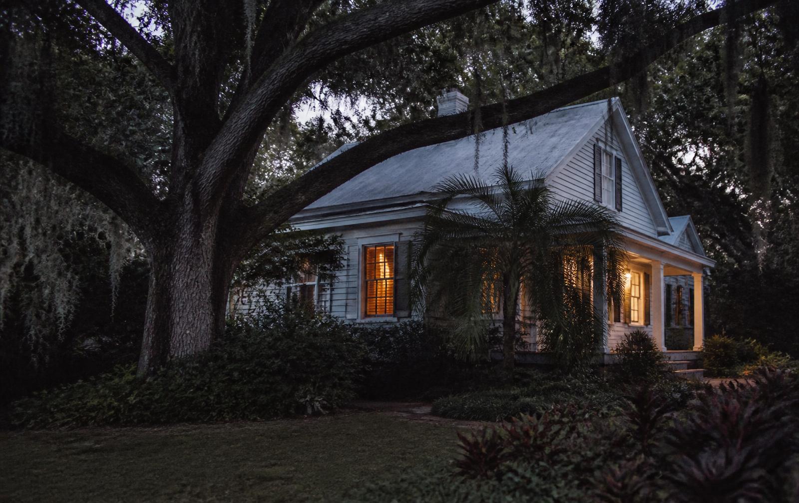 An older Florida home at dusk, seen through oak branches, with one window lit